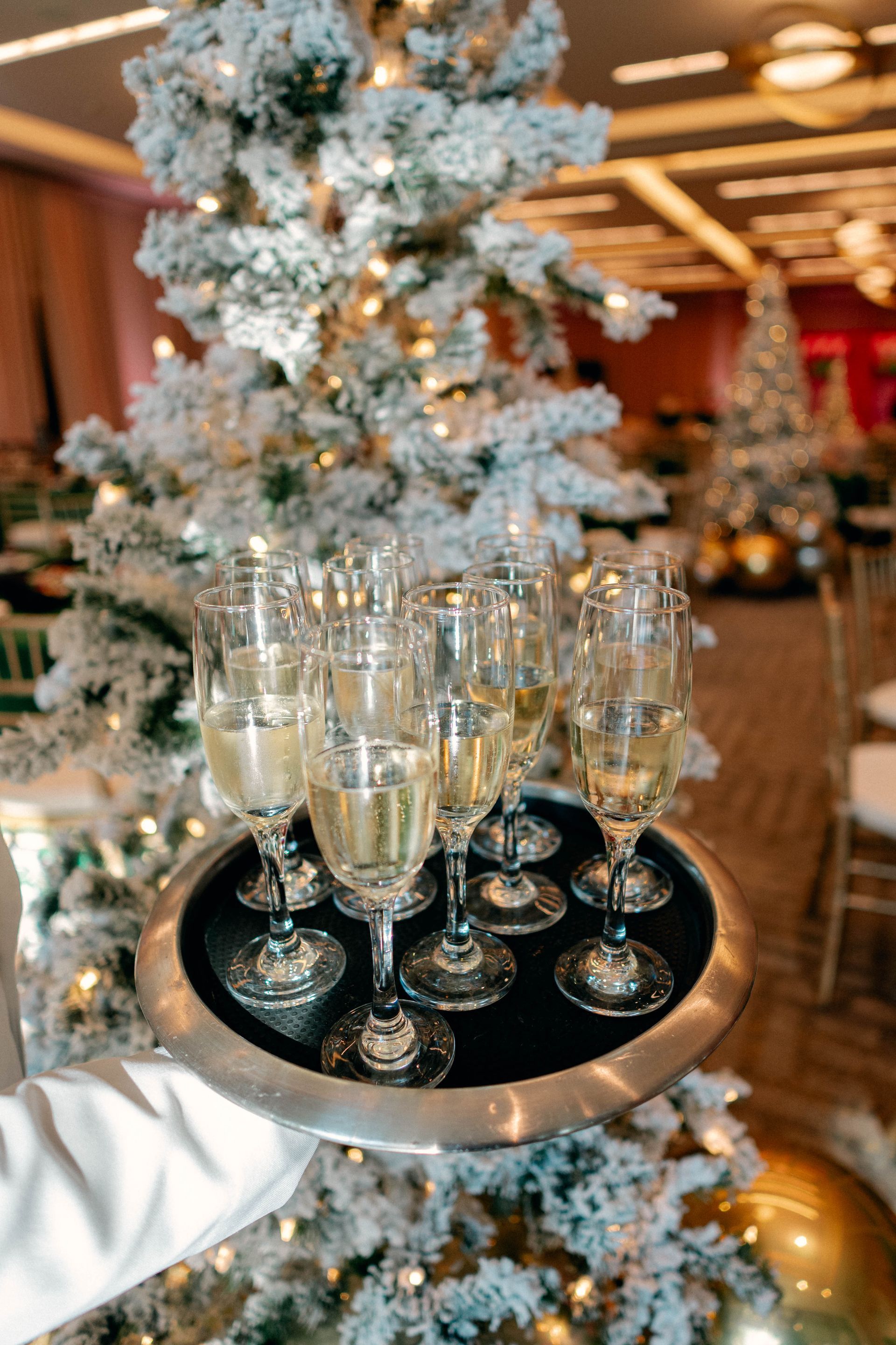 A server holds a tray of champagne flutes in a festive setting with a snow-covered tree.