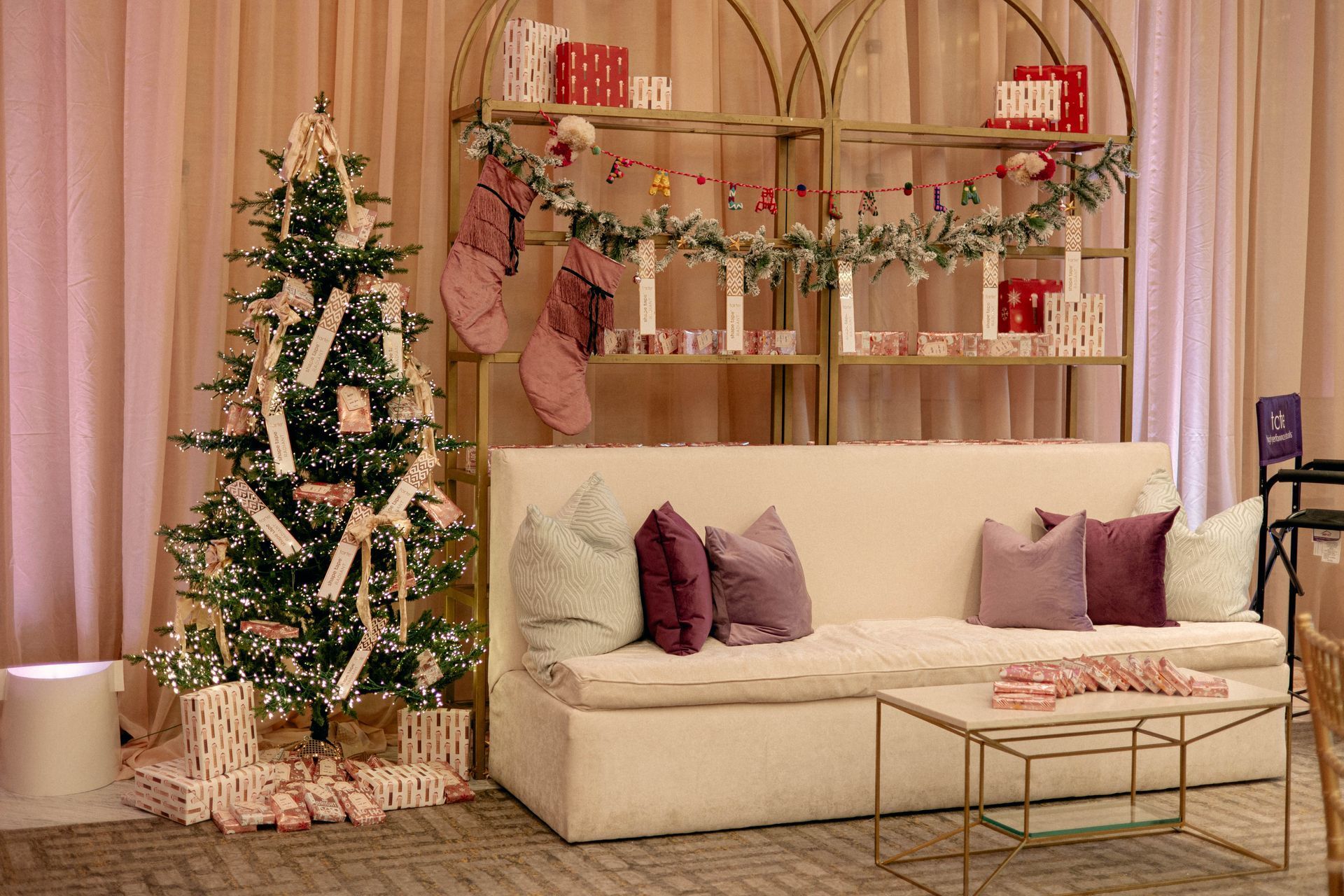 Christmas scene with decorated tree, sofa, and shelves filled with gifts. Soft pink and gold tones.