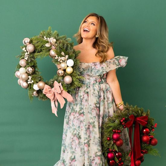 Woman in floral dress holds two Christmas wreaths against a green backdrop.