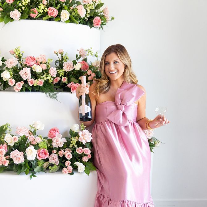 Woman in pink dress with champagne near flower-decorated cake. Smiling, celebratory pose.