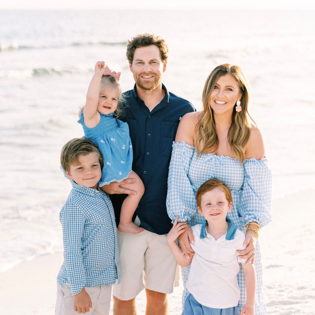 Family of five at beach, smiling. Two boys, a girl held by father, mother in blue gingham dress.