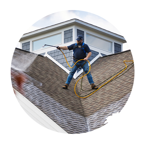 Technician cleaning a shingled roof with a pressure washer and safety harness