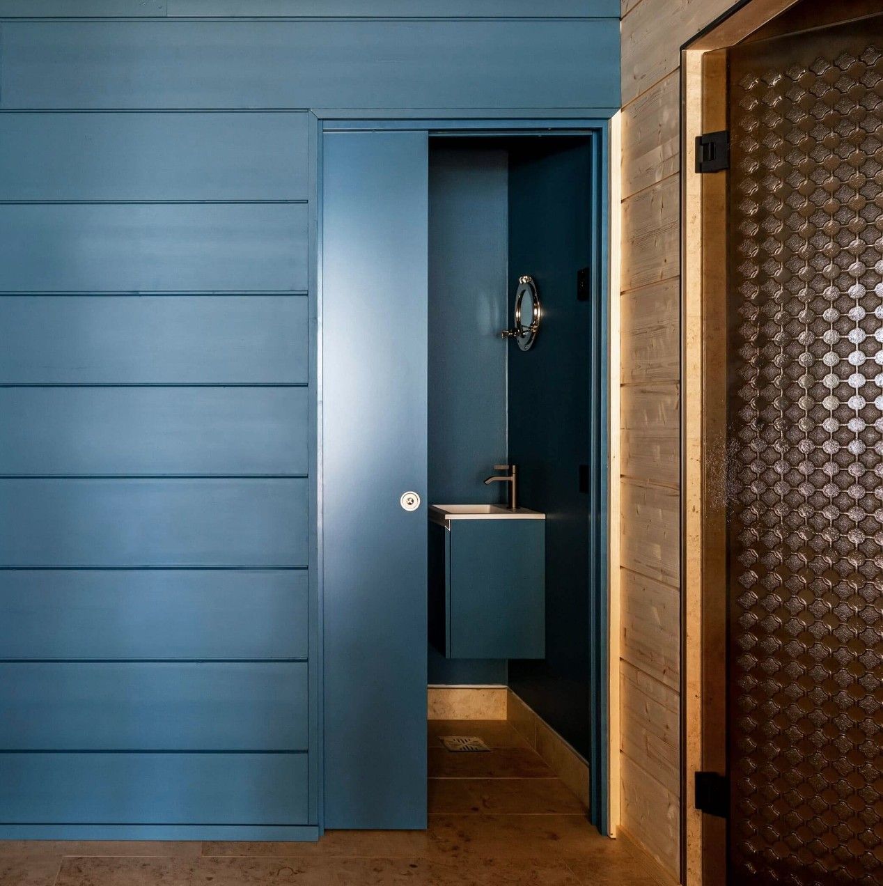 Parete rivestita di pannelli blu con una stretta porta aperta che rivela un piccolo bagno e pavimento in legno.