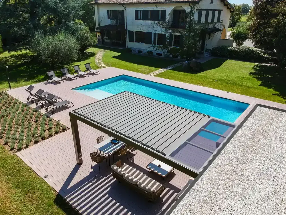 Piscina nel cortile sul retro di una casa a due piani, con pergola bioclimatica e sedie a sdraio intorno al terrazzo.