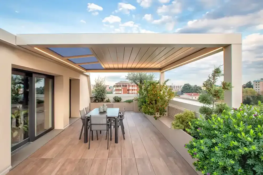 Patio moderno con tavolo da pranzo, pavimento in legno, pergolato e vegetazione lussureggiante con vista sulle case.