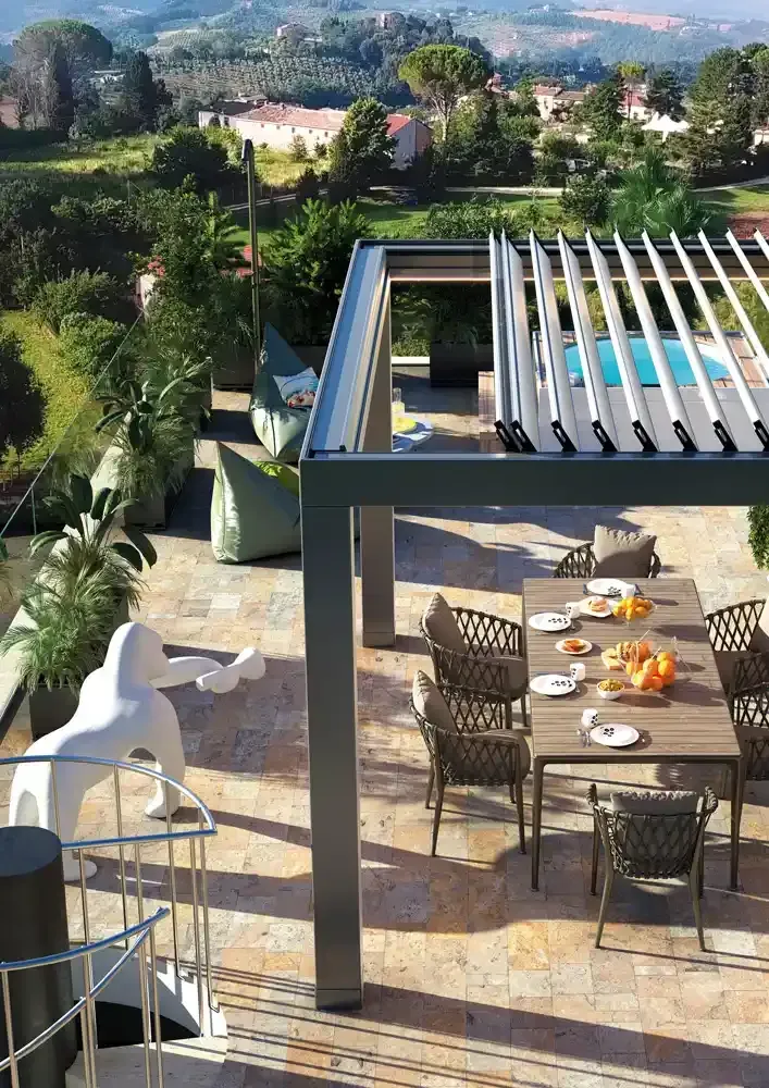 Pergola bioclimatica con tavolo, sedie e pergolato con vista su una verde collina e sulle case in lontananza.