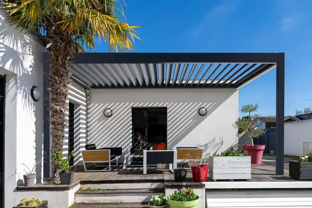 Patio moderno con pergola bioclimatica a righe, sedie a sdraio e piante in vaso all'esterno di una casa bianca.