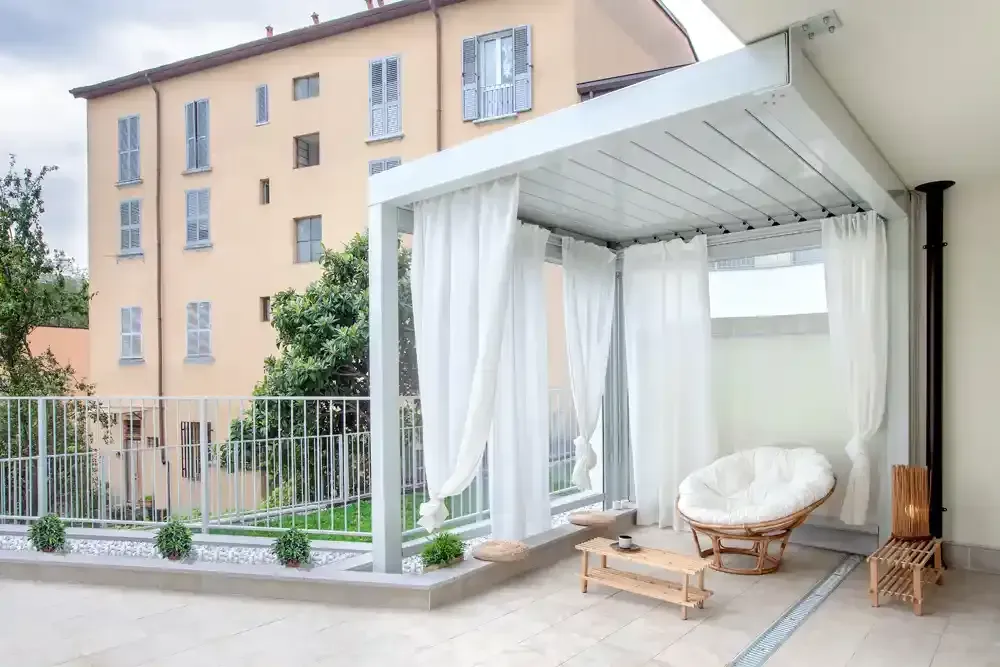 Pergola bioclimatica con tende, seduta in vimini e un tavolino accanto a un condominio beige.