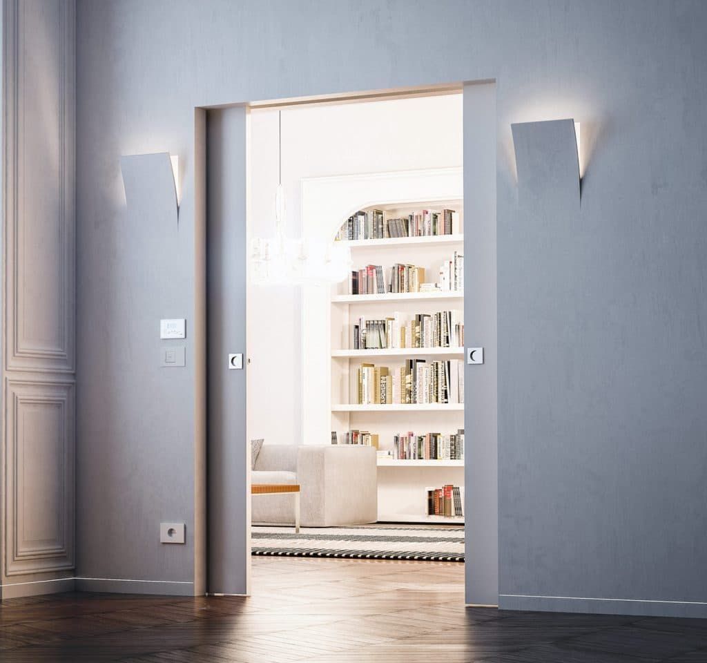 Si apre una porta su una stanza luminosa con librerie bianche e un divano, incorniciata da pareti grigie e pavimento in legno.