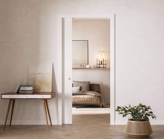 Camera da letto luminosa e minimalista con porta, piccola scrivania e pianta in vaso su un pavimento in legno chiaro.