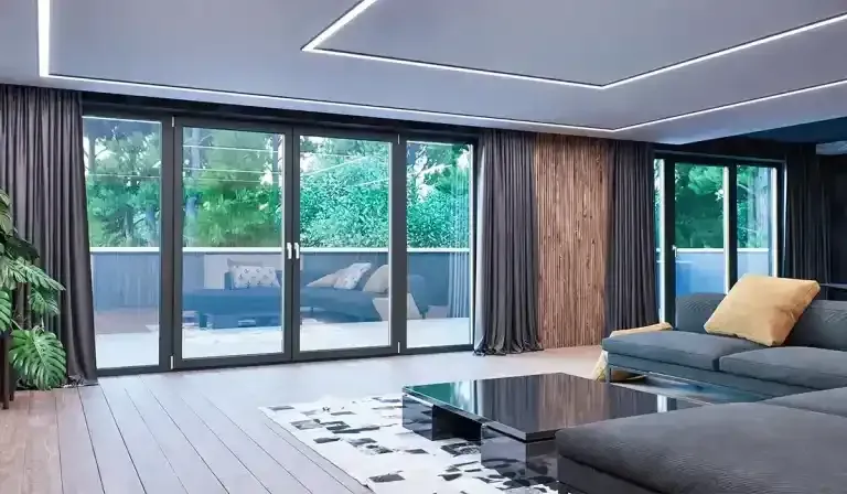 Soggiorno moderno con divano angolare grigio, dettagli in legno e ampie porte scorrevoli in vetro che si aprono su un patio e sul verde.