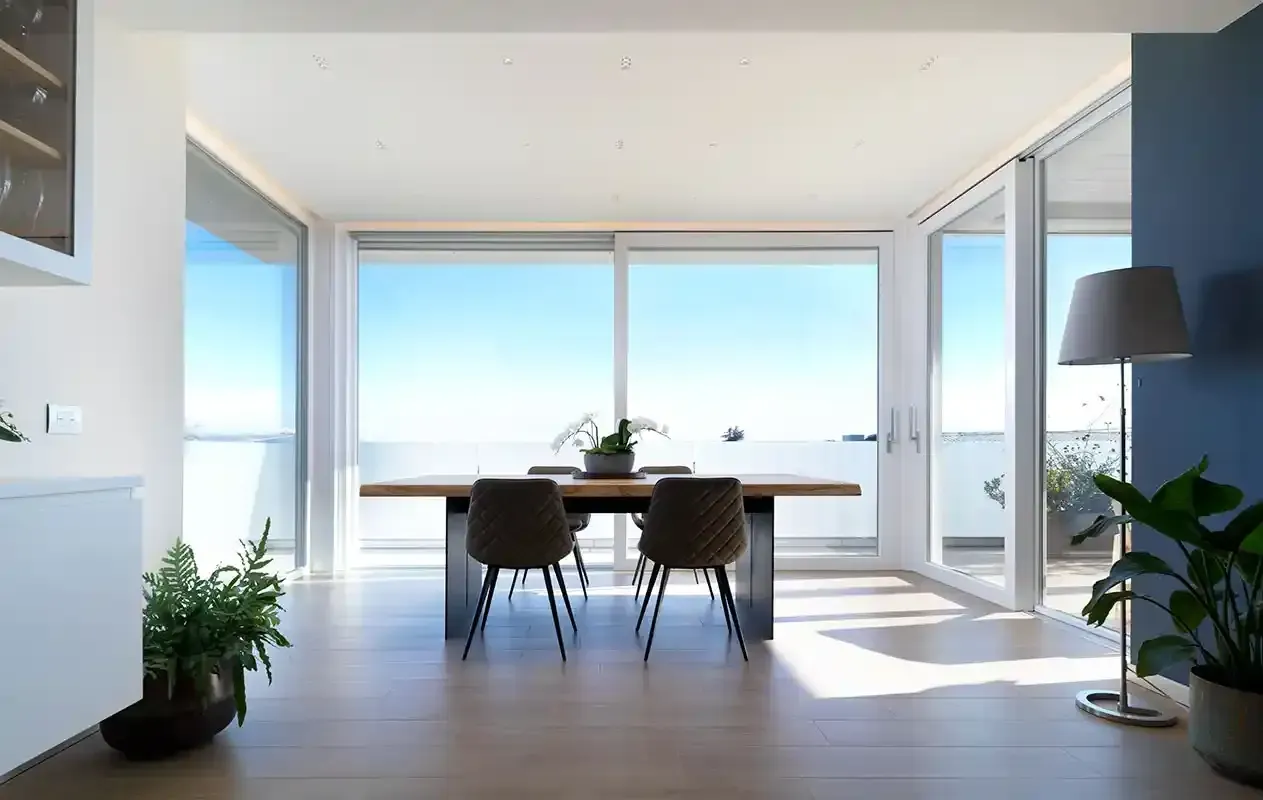 Luminosa sala da pranzo con ampie finestre, vista sull'oceano, tavolo in legno e piante in un interno moderno e minimalista.