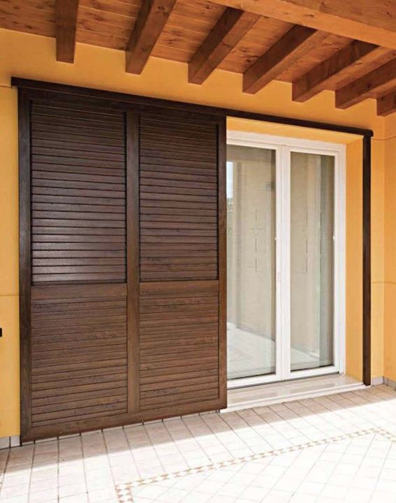 Persiane scorrevoli in legno accanto a una porta a vetri bianca su un patio coperto con travi a vista.
