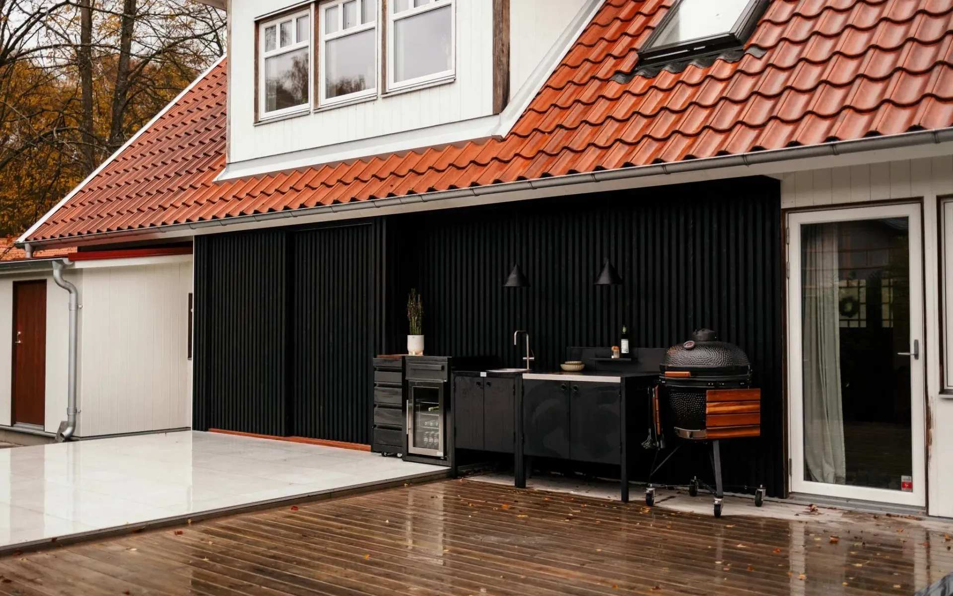 Esterno della casa con rivestimento nero, tegole rosse sul tetto e patio con mobili da esterno e barbecue.