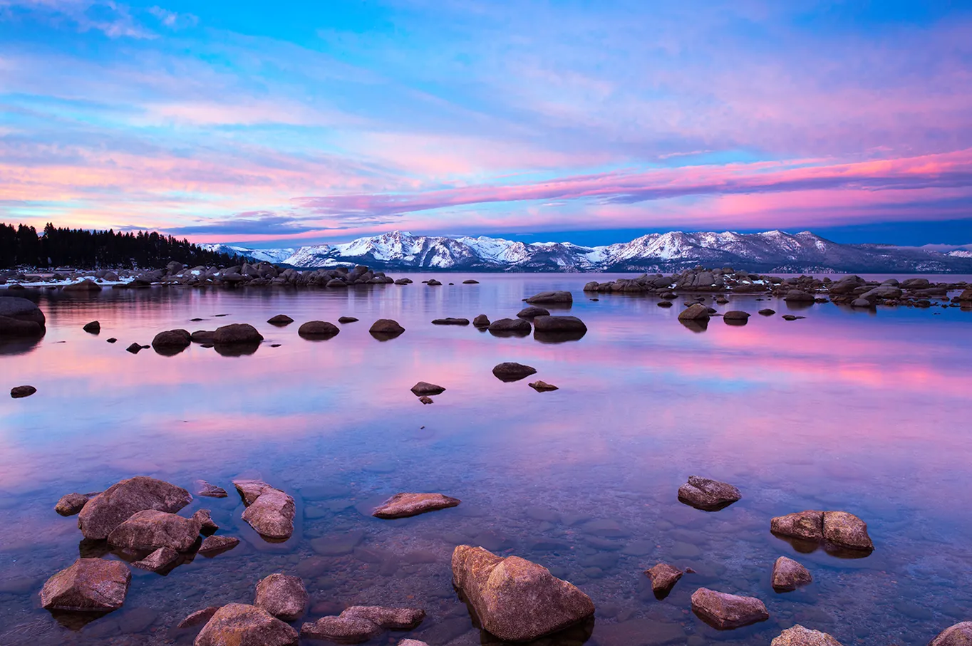 Snowy mountains reflected in a pink-blue lake with rocks in the foreground at sunset