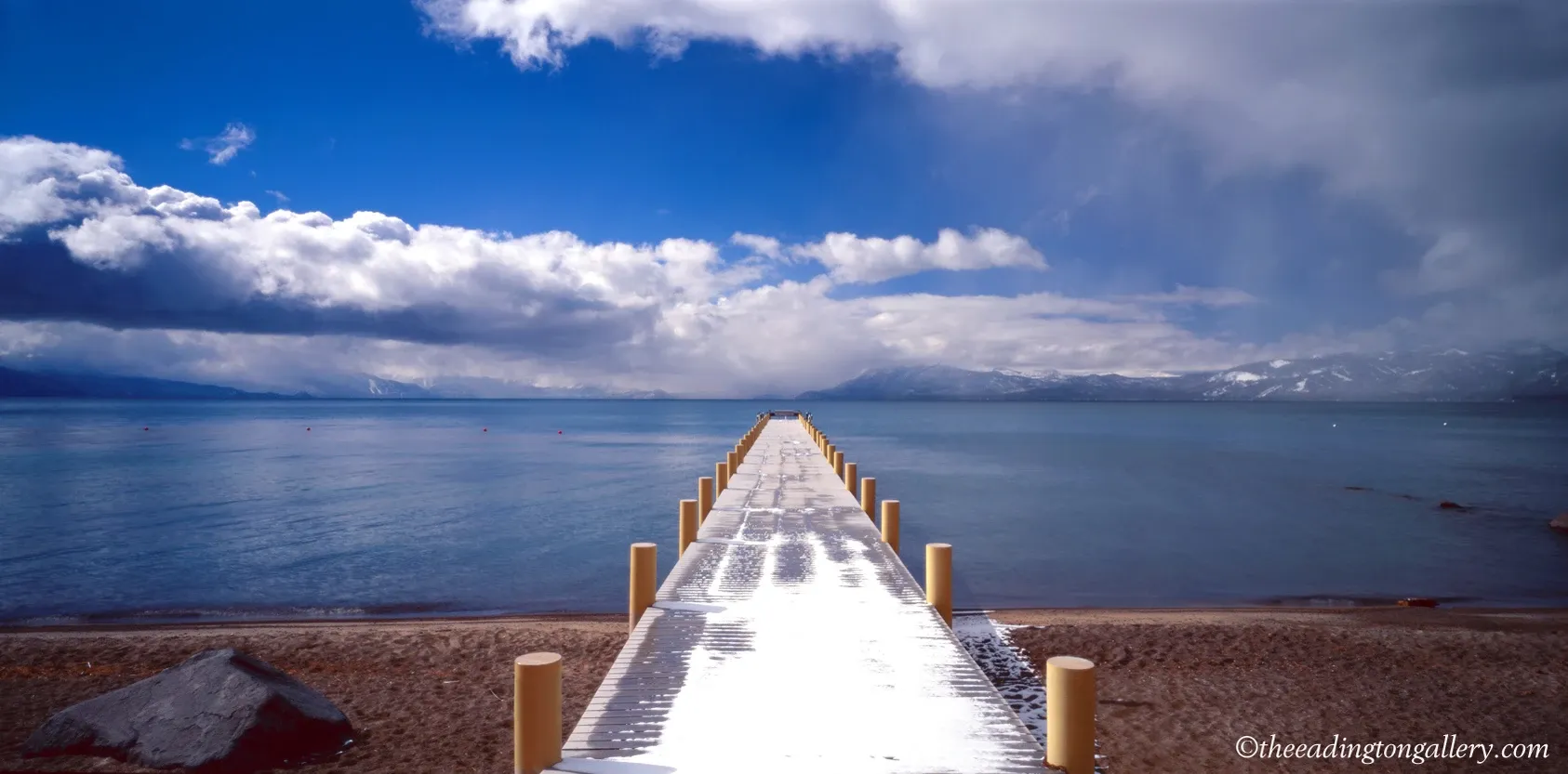 Snow-covered pier extends into a calm blue ocean under a bright cloudy sky.