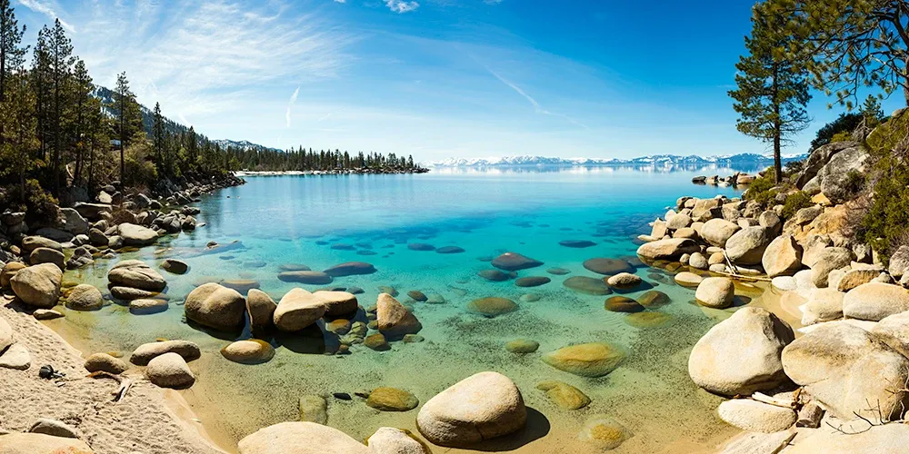Clear turquoise lake with rocky shoreline, pine trees, and distant mountains under a blue sky