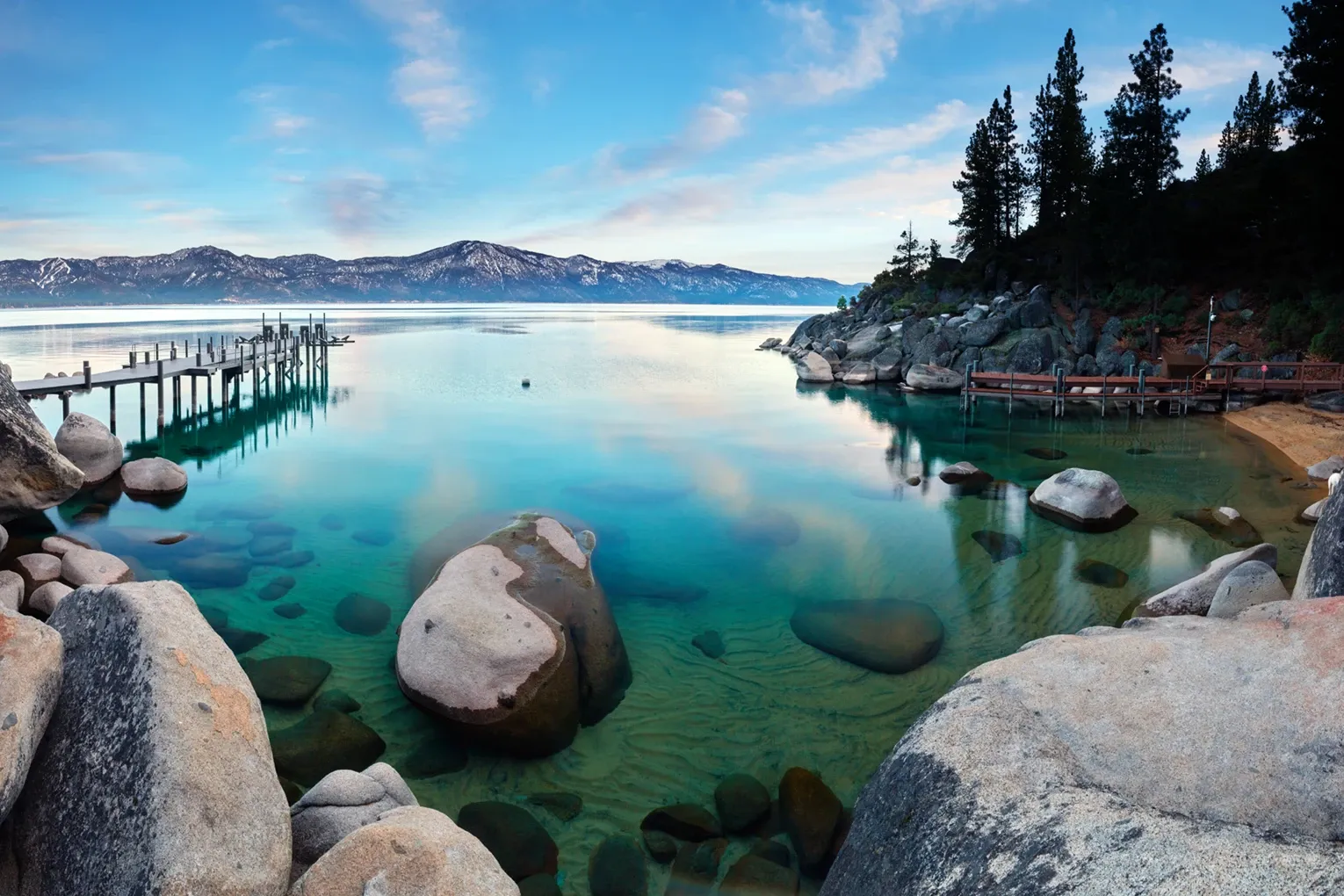 Rocky lakeshore with clear turquoise water, dock, and pine trees under a bright blue sky