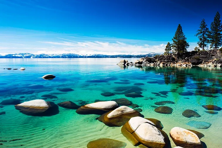 Clear turquoise lake with rocks in the foreground and snow-capped mountains under a blue sky
