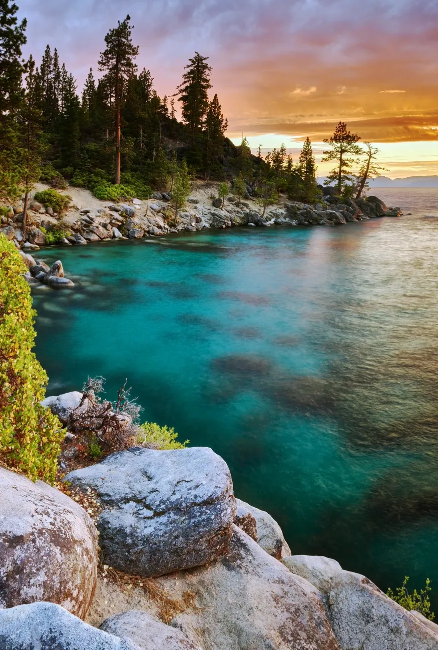 Turquoise water beside rocky pine shore at sunset, with glowing sky and calm bay