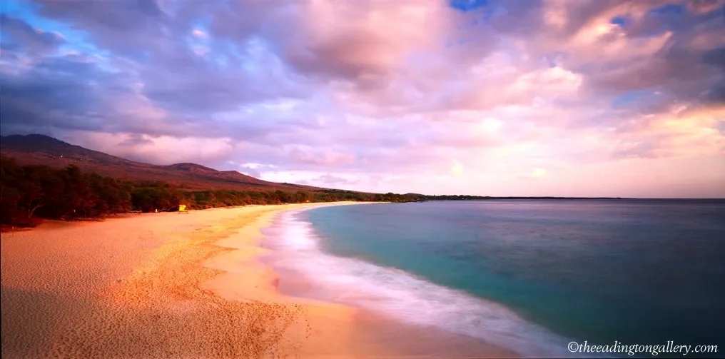 Sunset over a sandy beach with gentle waves, pink clouds, and distant hills
