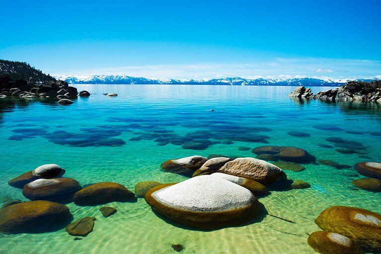 Rocky shoreline with clear turquoise water, smooth stones, and distant snowy mountains under a bright blue sky