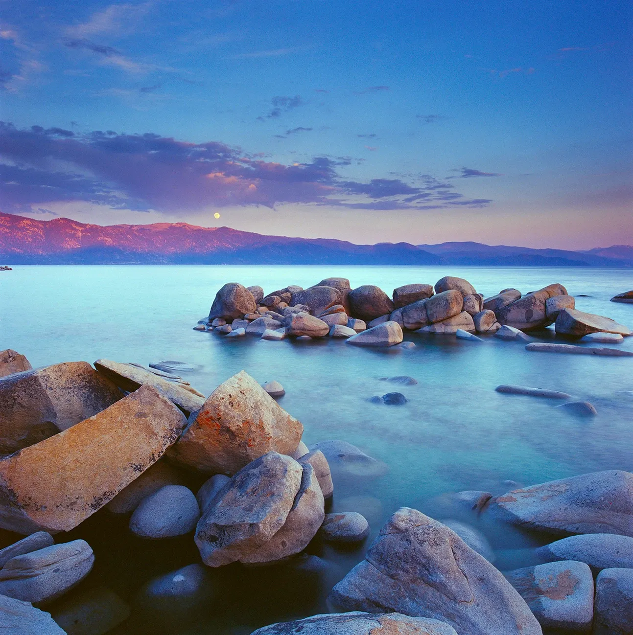 Rocky shoreline at dusk with calm blue water and distant purple mountains under a colorful sky