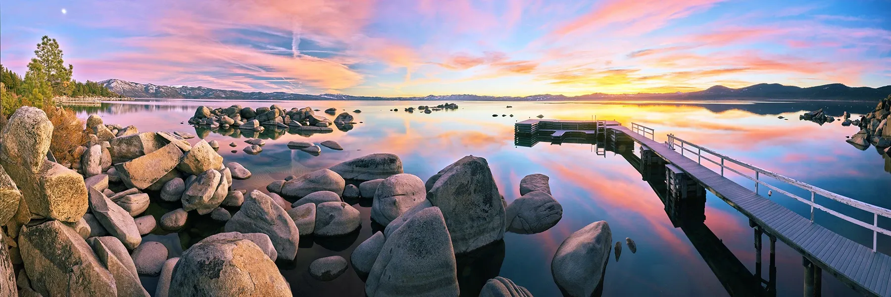 Rocky shoreline at sunset with calm water reflecting pink and orange sky, and a long pier extending out