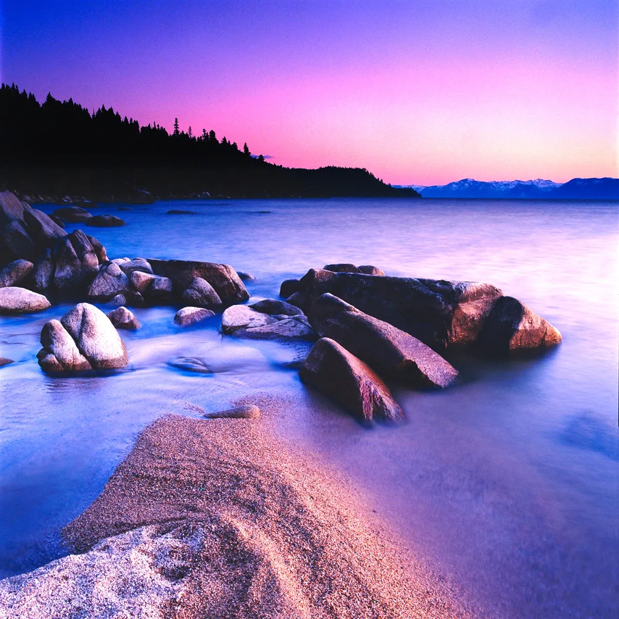 Rocky shoreline at dusk with calm water, distant hills, and a pink-purple sky