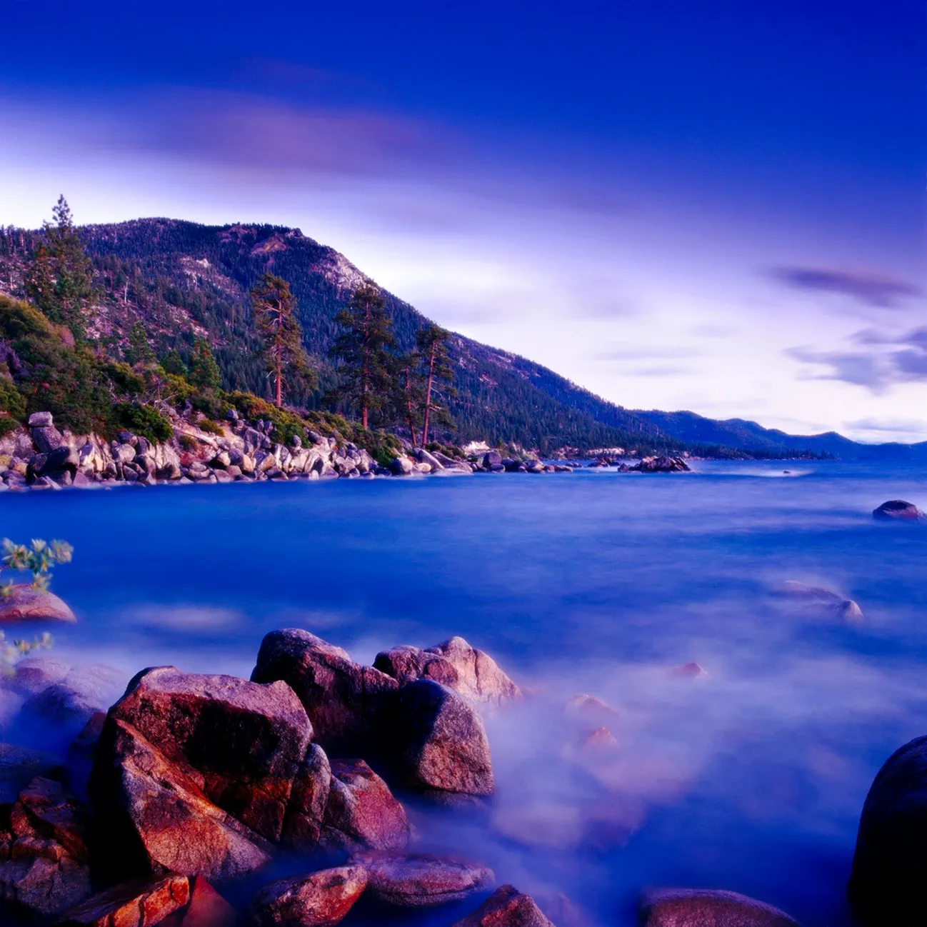 Rocky coastline with calm blue water, forested hills, and a purple twilight sky