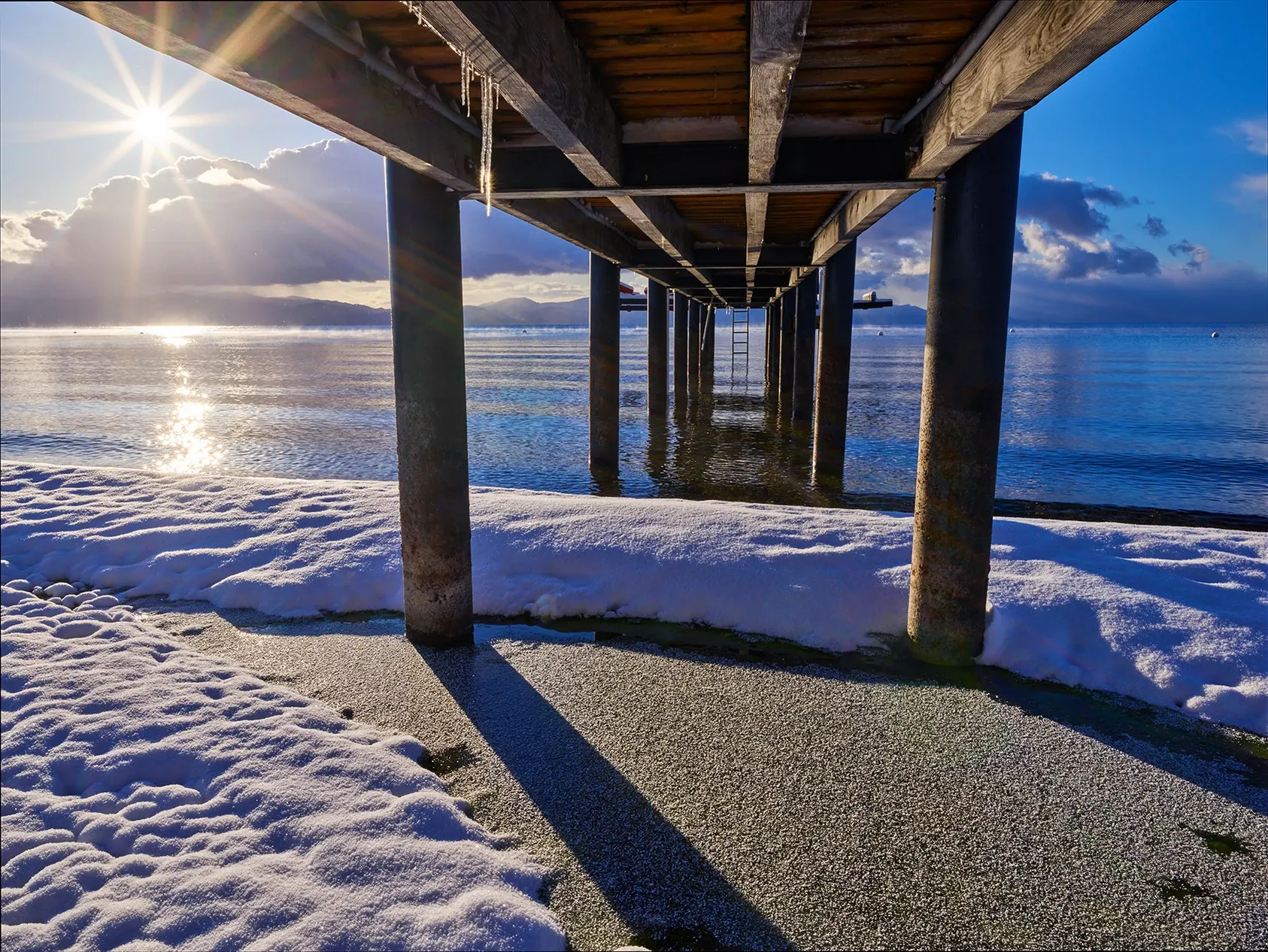 Sunlit snowy pier over a calm blue sea with long shadows and a bright sky