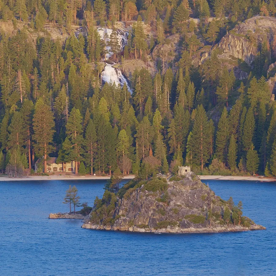 Small rocky island in a blue lake, backed by pine forest and a waterfall on a mountain slope.