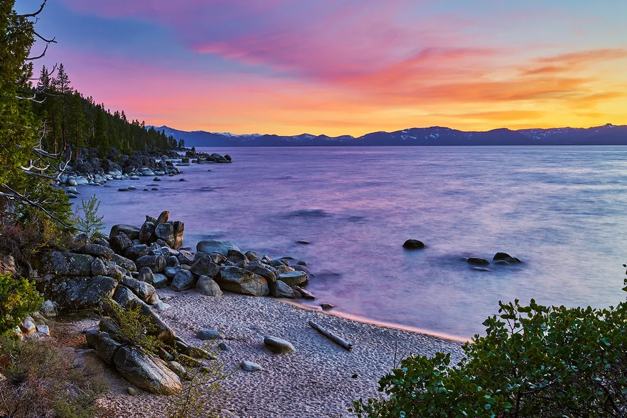 Rocky lakeshore at sunset with calm water and a pink-orange sky