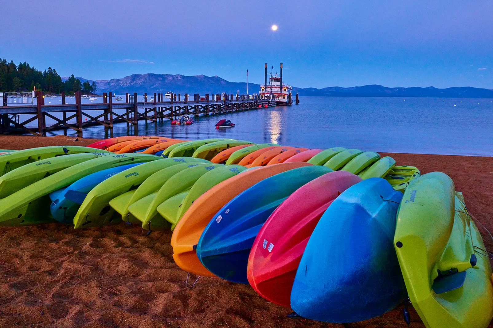 Colorful kayaks lined up on a sandy beach beside calm water at dusk, with a dock and moonlit sky.