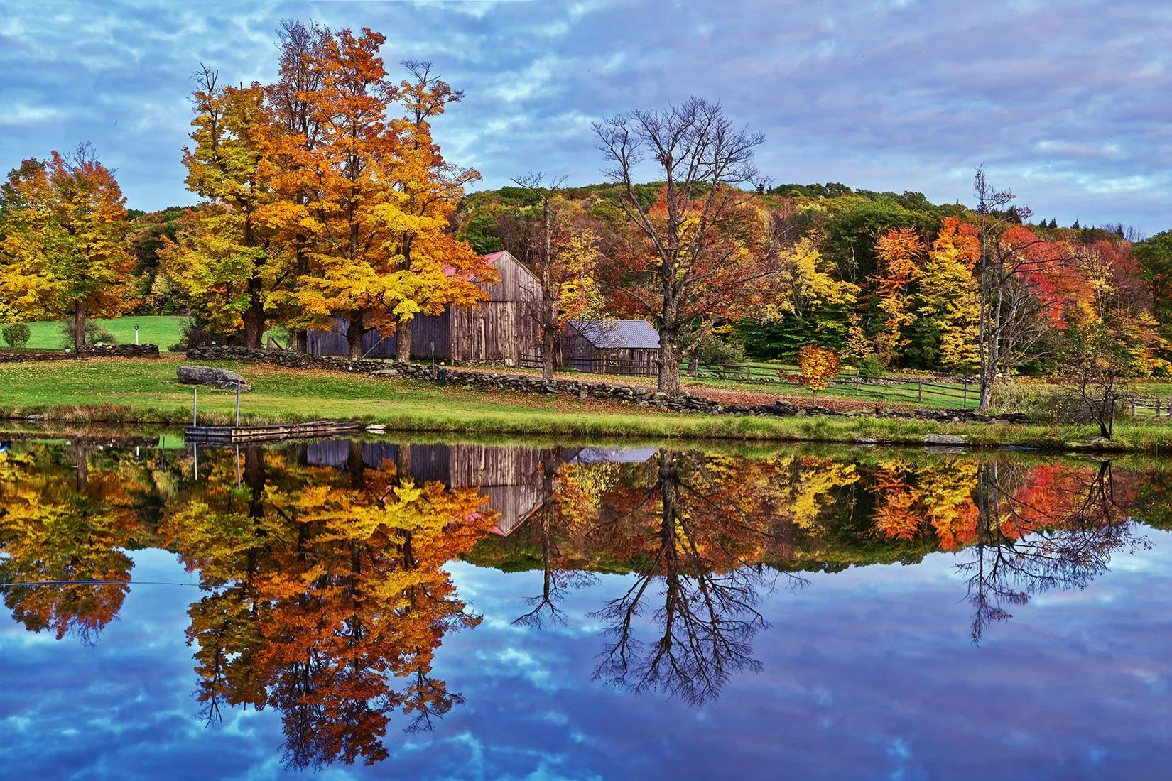 Autumn trees and a farmhouse reflected in a calm pond under a clear blue sky