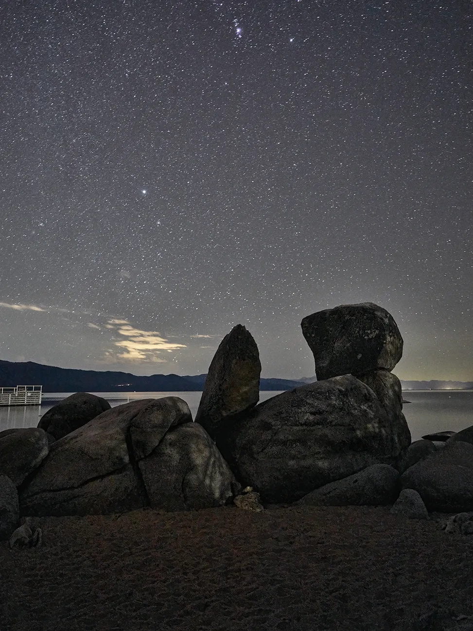 Starry night sky over silhouetted coastal rocks and a faint city glow on the horizon