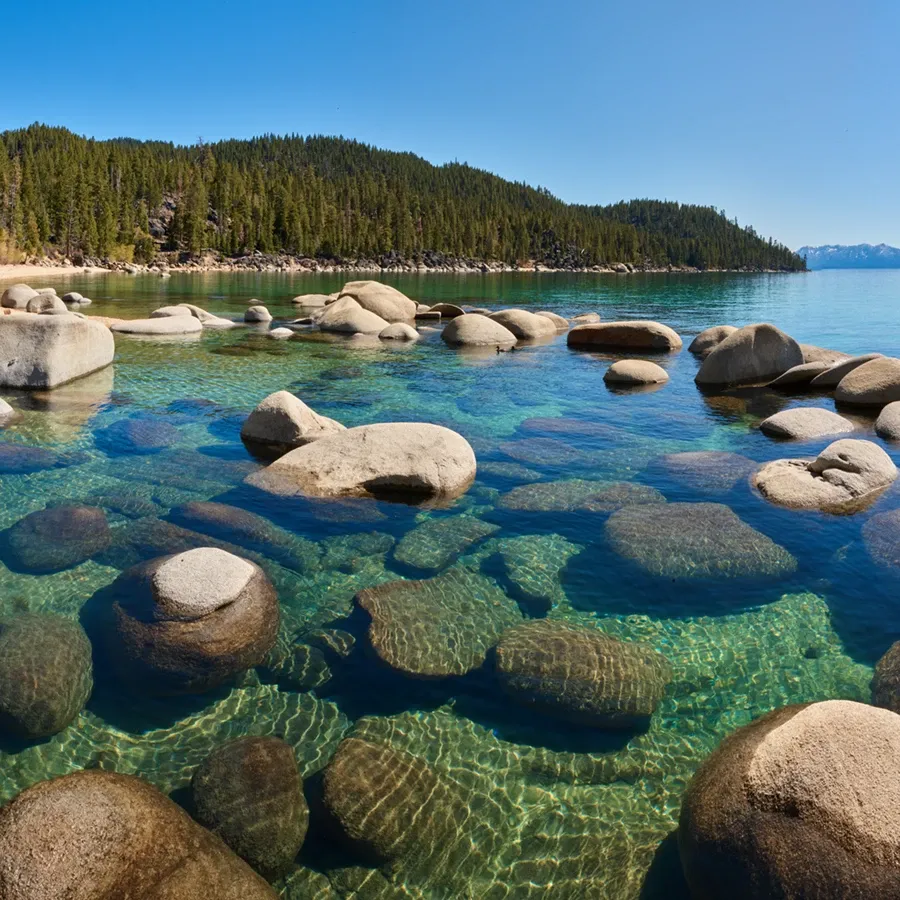 Clear turquoise lake with smooth boulders and a forested shoreline under a blue sky