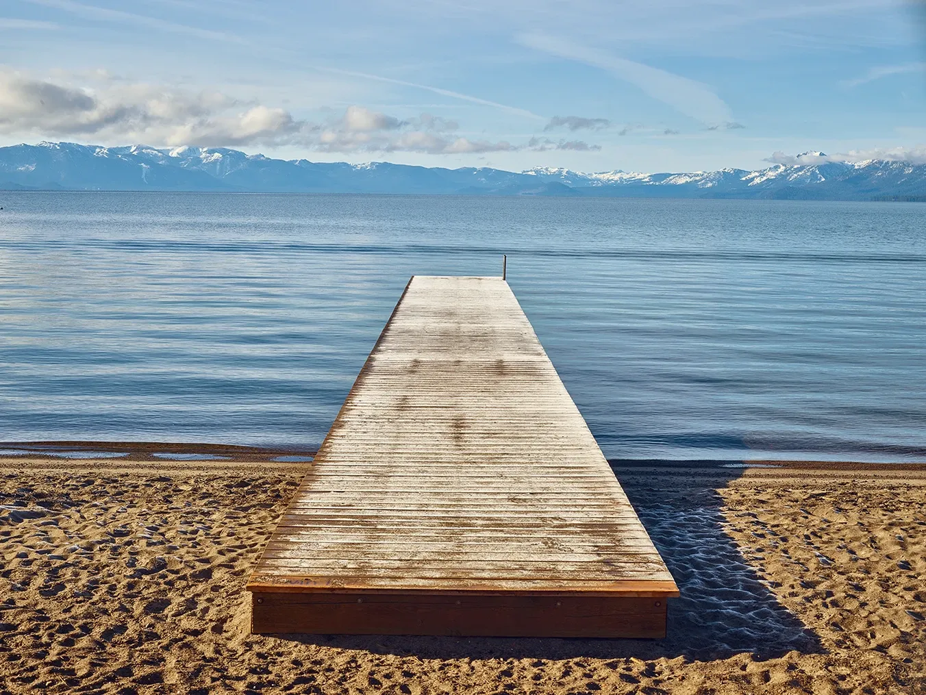 Wooden dock extending into a calm lake with snowy mountains in the distance under a blue sky