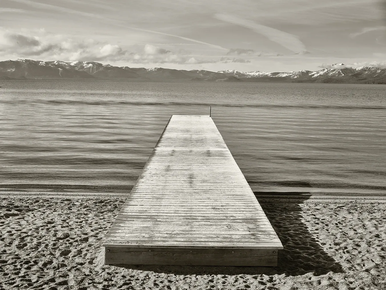 Wooden dock extends into a calm lake under a cloudy sky, with pebbled shore in the foreground.