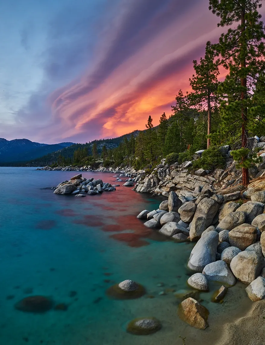Rocky turquoise shoreline at sunset with pink-purple clouds and pine trees