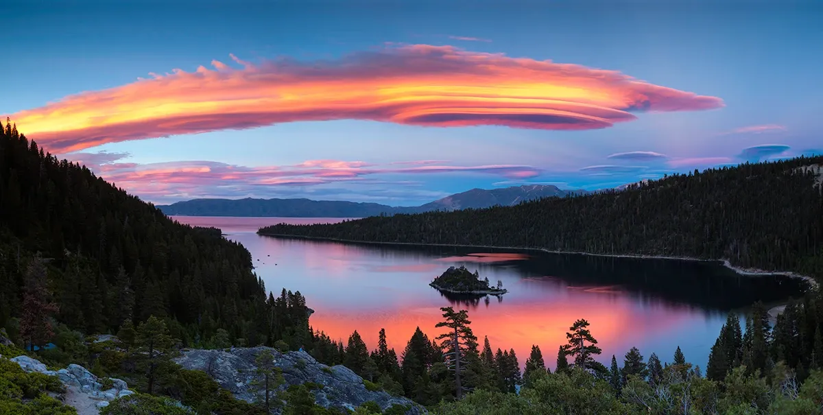 Lone island on a calm lake at sunset, with pink clouds and dark forested hills.