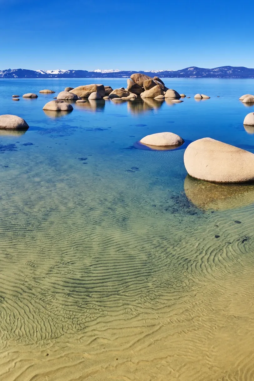 Clear blue lake with sandy shallows and rounded rocks under a bright sky, with distant mountains on the horizon