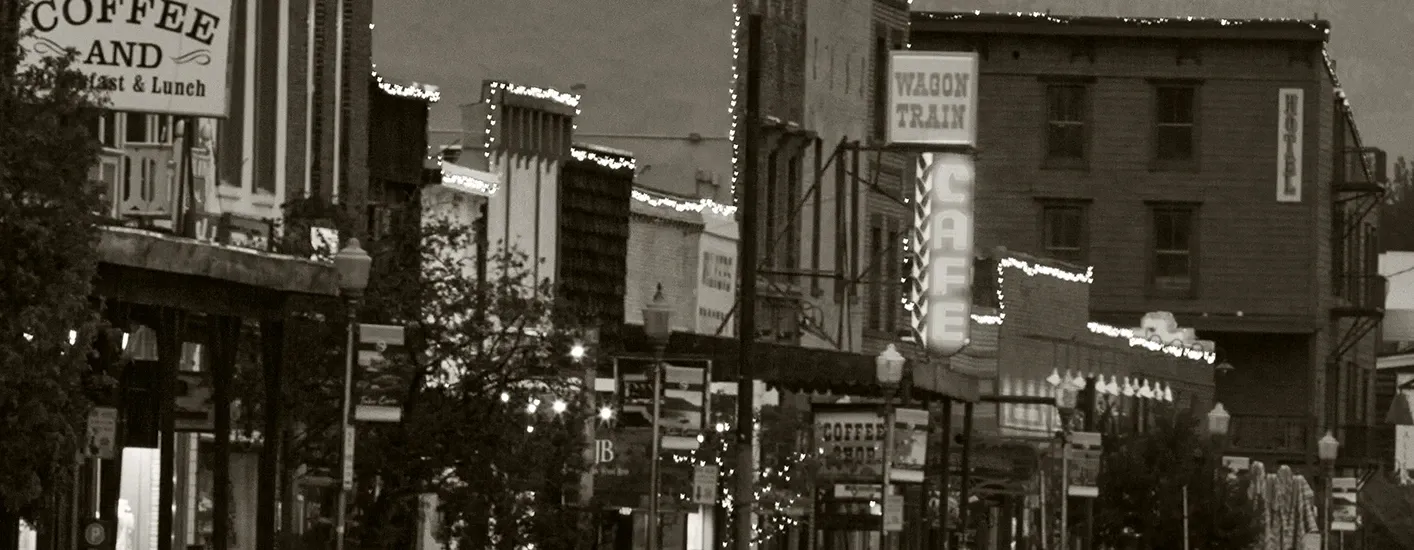 Sepia-toned main street with storefronts, hanging signs, and parked cars in a small town.