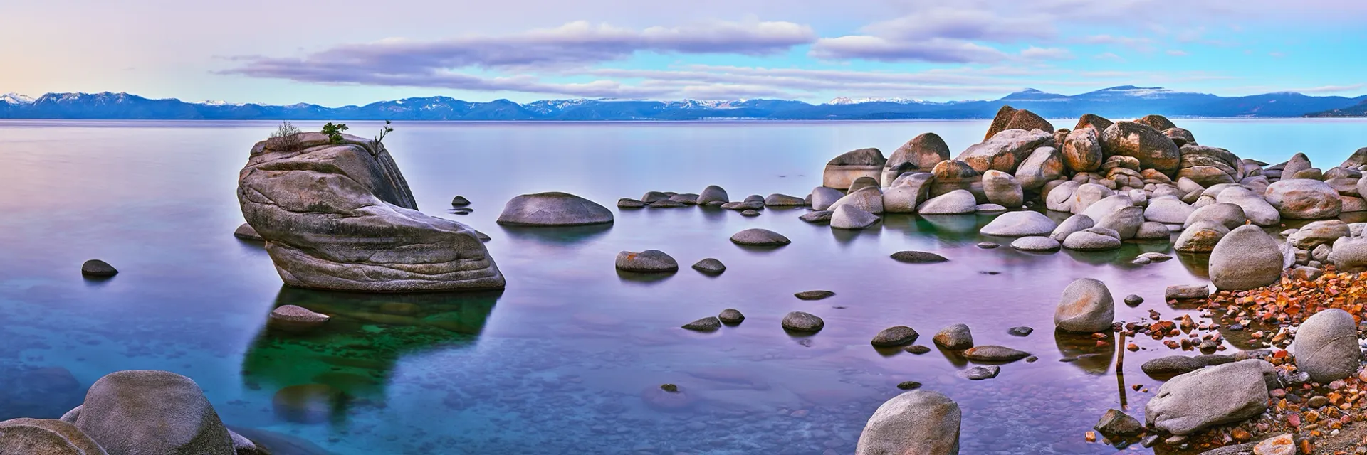Rocky lakeshore at dusk with calm water, scattered boulders, and a distant blue horizon