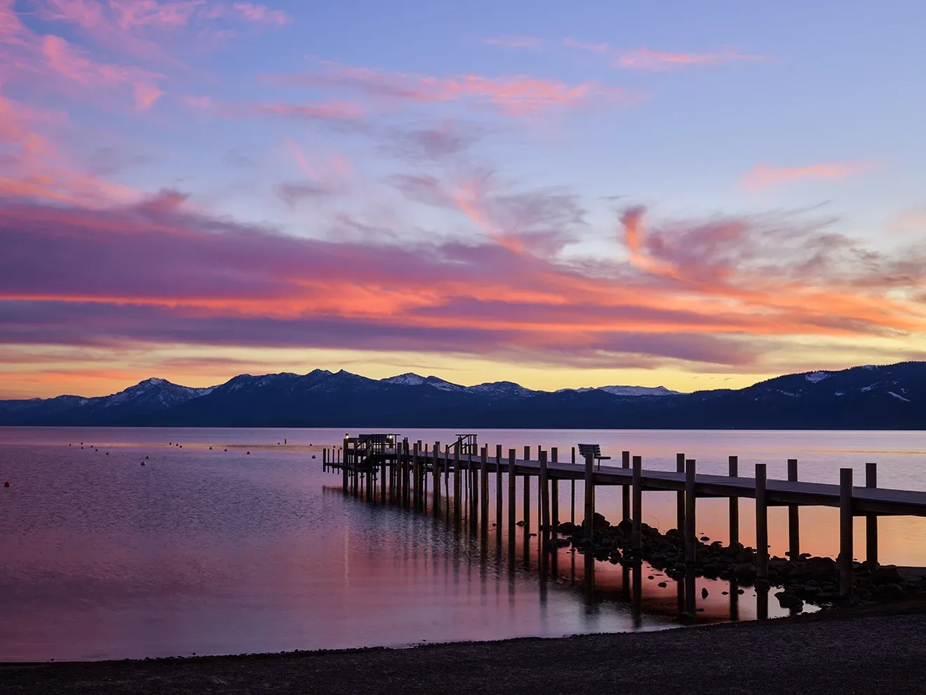 Sunset over calm water with a wooden pier and silhouetted hills, pink and orange sky reflected on the lake