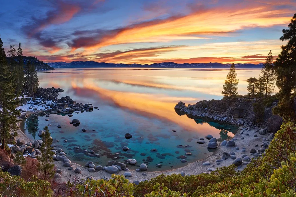 Sunset over a turquoise lake with rocky shores, pine trees, and colorful clouds reflected in calm water