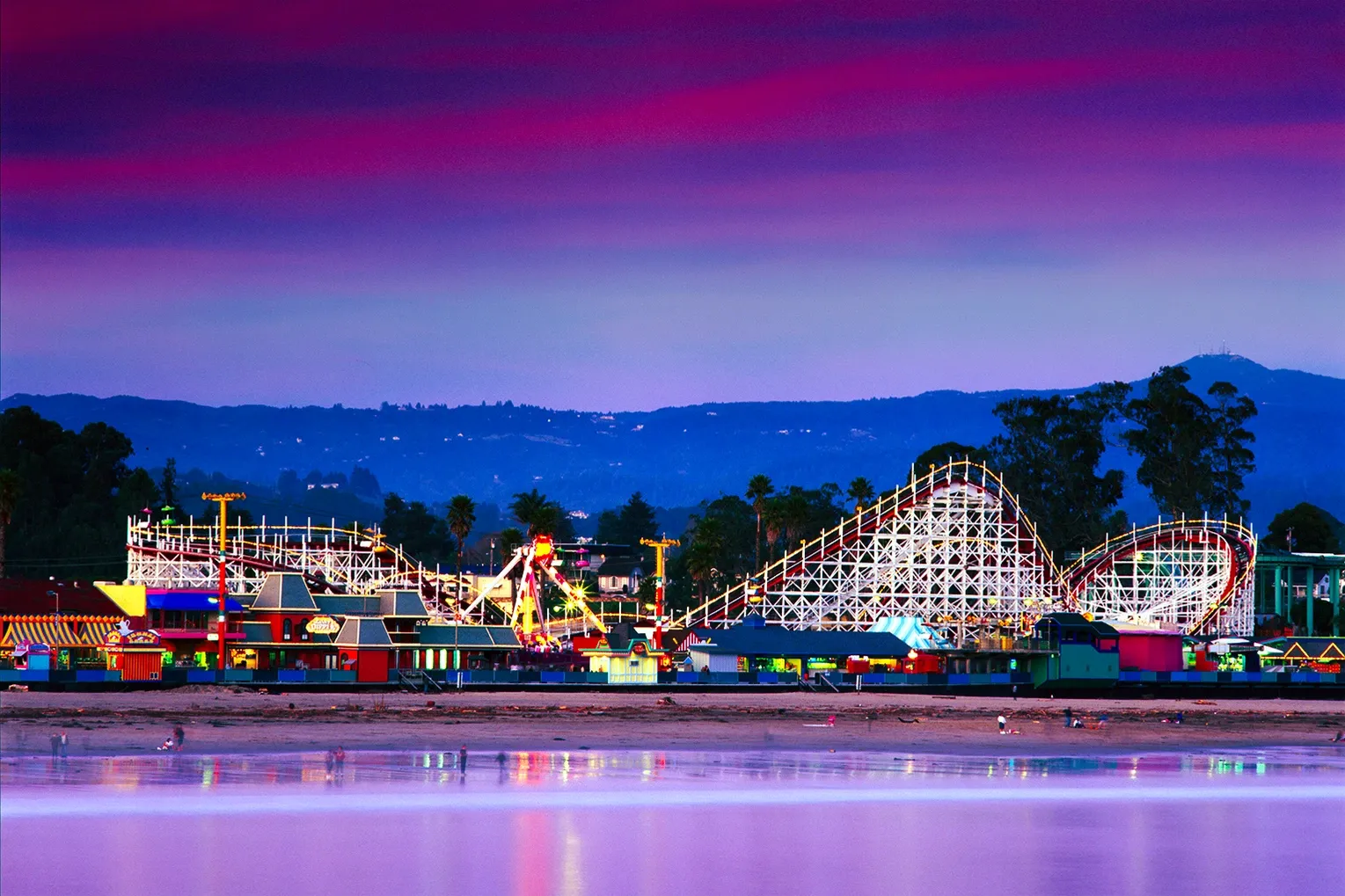 Colorful amusement park at dusk with a roller coaster reflected in calm water beneath a pink-purple sky