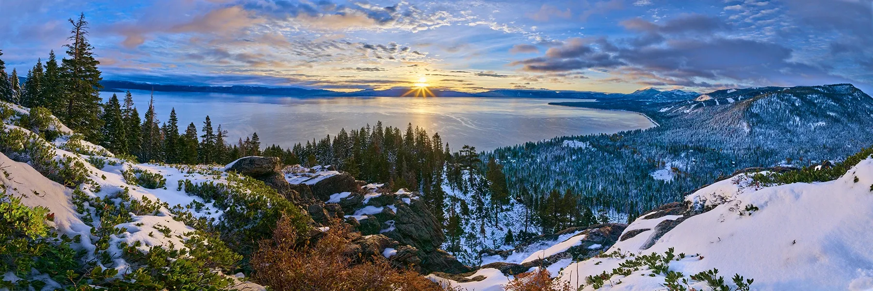 Snowy mountain landscape at sunrise with pine trees, distant peaks, and a glowing sky over a frozen valley