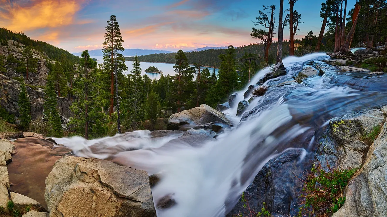 Mountain waterfall rushing over rocks at sunset, with pine trees and a lake in the distance