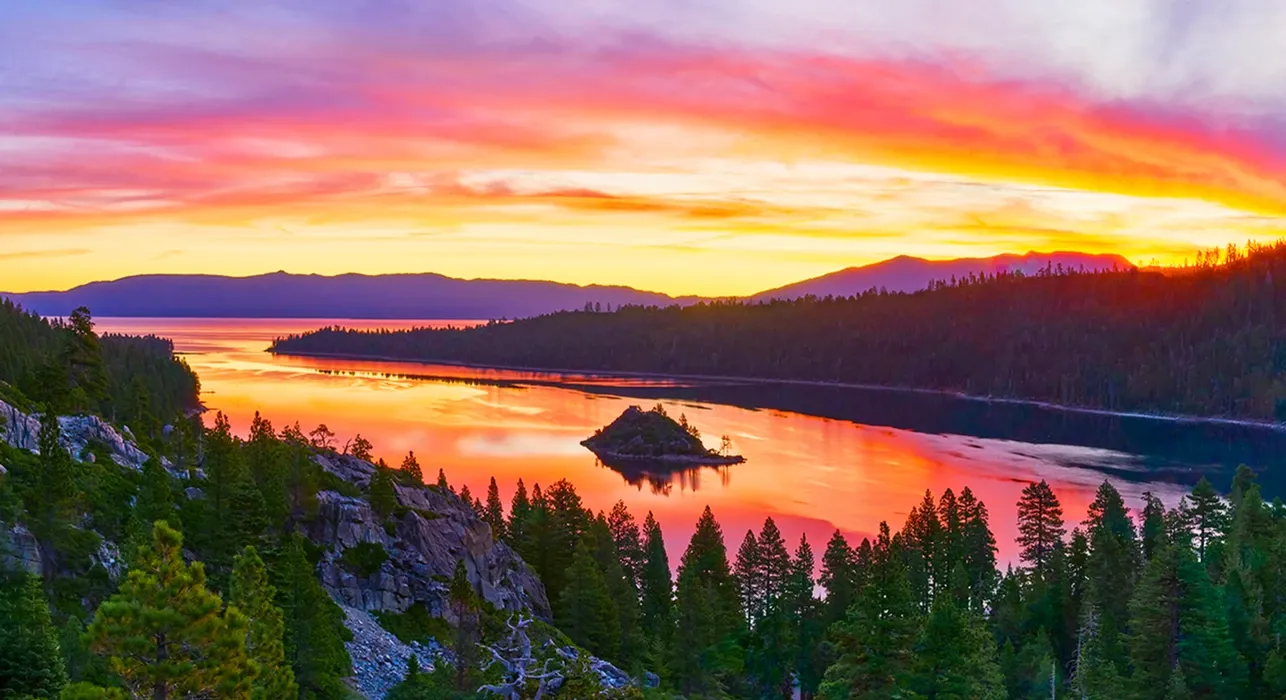 Vibrant sunset over a calm lake with a small island, pine forest, and colorful clouds
